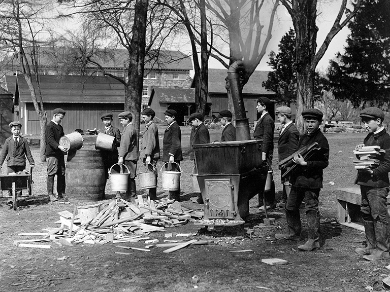 About 10 young boys line up with buckets to pour their sap into a barrel while another 2 young boys carry wood to feed the fire of the wood stove featured in the center.  1 other young boy hauls wood in a wheel barrel.  All the boys are out in a wooden field wearing boots, jackets and caps.
